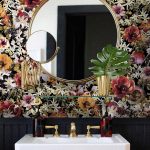 Kúpeľňa s výraznou kvetinovou tapetou na stene, na nej visí okrúhle zlaté zrkadlo a presklená polička, pod nimi je biele umývadlo so zlatou batériou