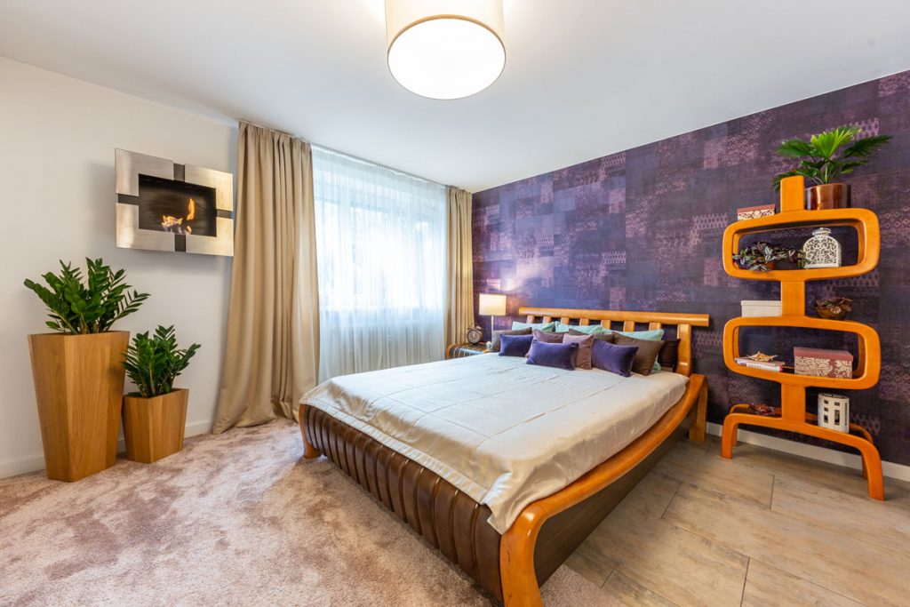spálňa s fialovou vzorovanou tapetou, zariadená exotickým nábytkom z Indonézie, s masívnou posteľou a otvorenou skrinkou z ohýbaného lakovaného dreva, masívnymi drevenými kvetináčmi a krbom zaveseným na stene