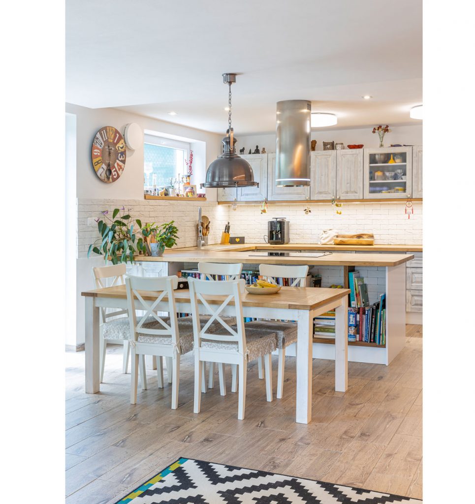 kuchyňa vo vidieckom štýle s industriálnymi prvkami, drevený stôl so stoličkami, s industriálnym lustrom, ostrovček s úložným priestorom na knihy a kuchynská linka v bielej farbe s tehlovým obkladom na stene