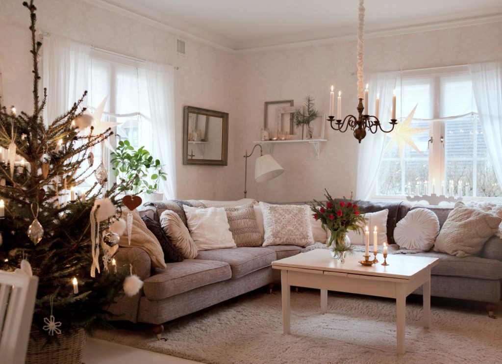vianočne zariadená obývačka s rohovým gaučom v neutrálnej farbe, na ktorom je veľa vankúšov v odtieňoch bielej a hnedej, s bielym stolíkom s vysokými svietnikmi a vázou s čečinou a kvetinami, mäkkým kobercom a vianočným stromčekom s prírodnými ozdobami