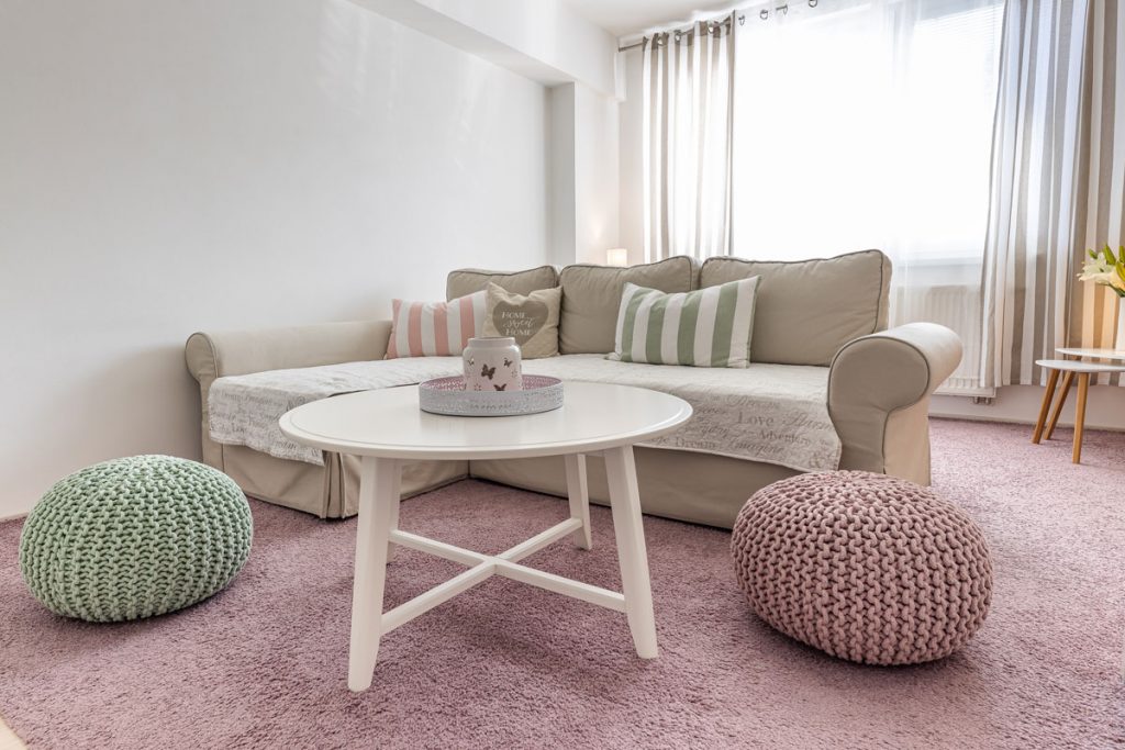 Obývačka v romantickom vidieckom štýle s pohovkou v neutrálnej farbe, pastelovým ružovým a zeleným pufom, ružovým kobercom a vidieckym okrúhlym stolíkom.