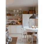 kuchyňa vo vidieckom škandinávskom štýle s bielym okrúhlym rozkladacím stolom, bielymi stoličkami, bielou kuchynskou linkou, pleteným prírodným behúňom a otvorenými policami, na ktorých sú poukladané riady a staré drevené truhlice