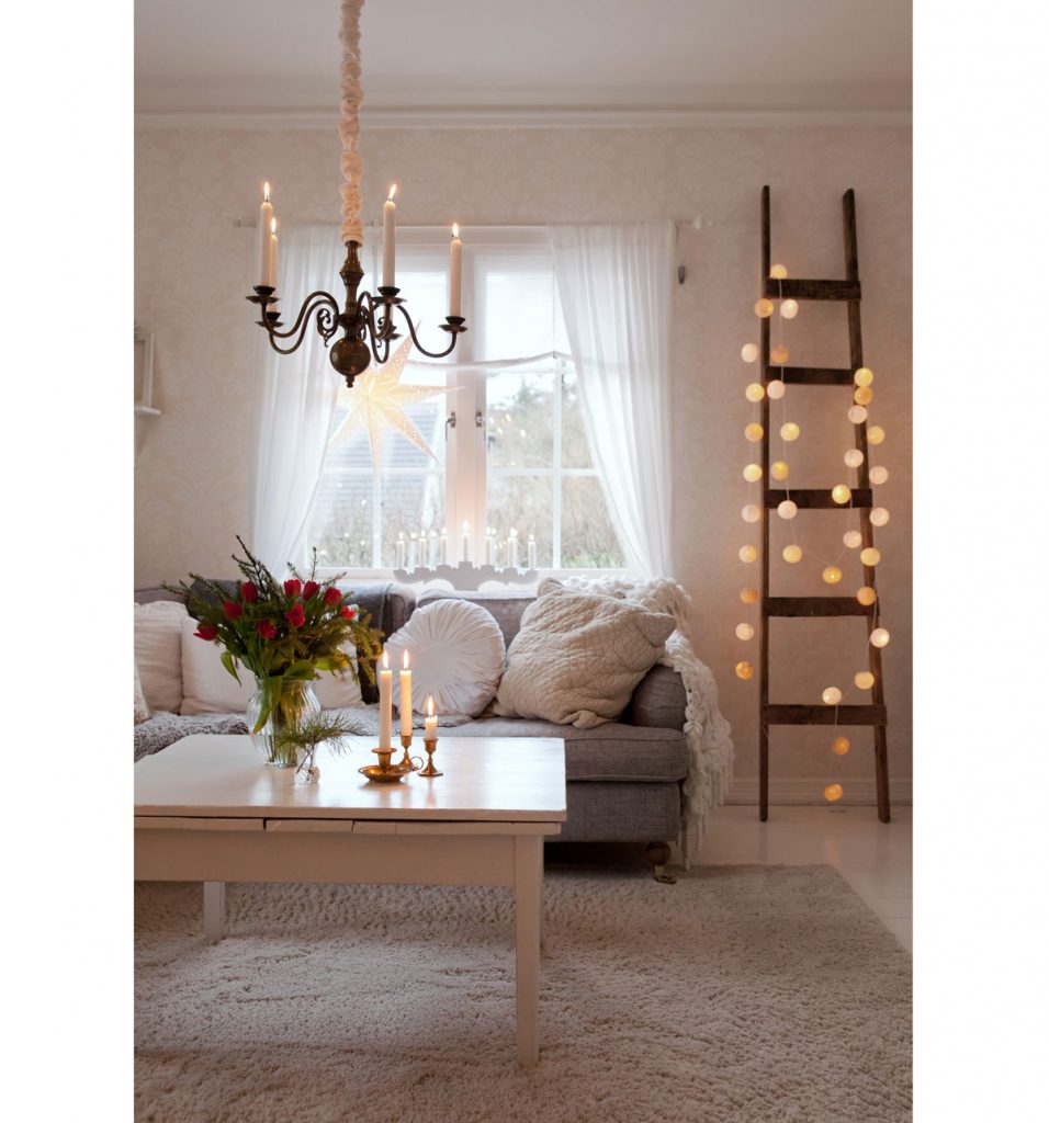 škandinávsky vidiecky interiér vyzdobený na Vianoce, so sedačkou a pohodlnými vankúšmi z prírodných materiálov, s bielym stolíkom, na ktorom sú stojany so sviečkami a váza so živými kvetmi