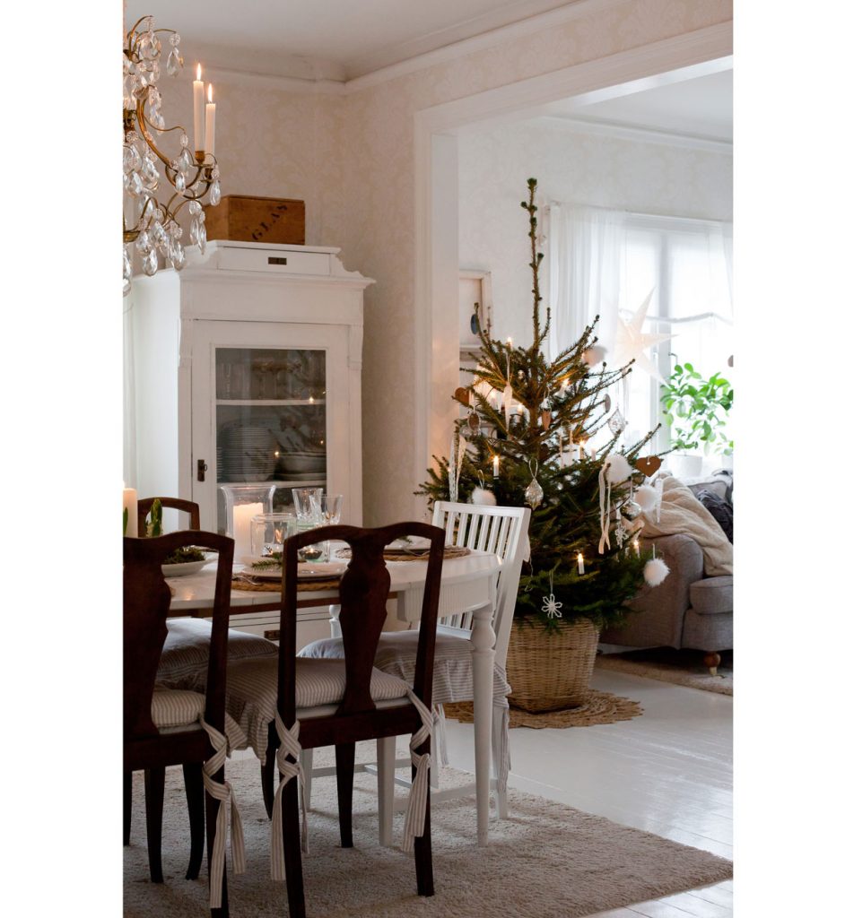 škandinávsky vidiecky interiér s bielym stolom, drevenými stoličkami, bielym kredencom, so sviatočným vianočným prestieraním a živým vianočným stromčekom v kvetináči