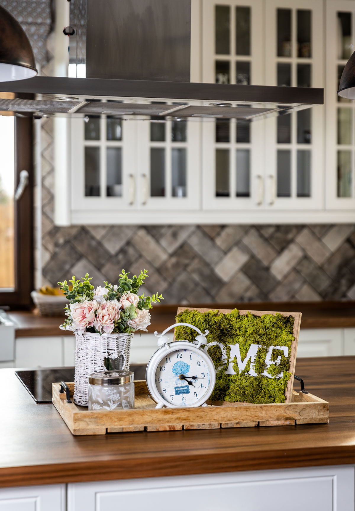 Kuchynská dekorácia z drevenej tácky, na ktorej je machový obraz s nápisom HOME, biely budík, sklenená nádoba a prútená nádoba s kvetmi.
