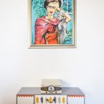 Interiér s obrazom Fridy Kahlo a farebnou komodou.