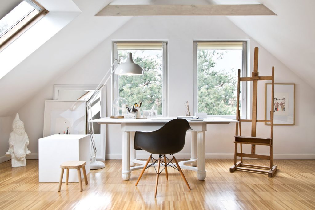 Minimalisticky zariadená pracovňa v podkroví drevenice, s bielym dizajnovým stolom, stoličkou a dreveným maliarskym stojanom.