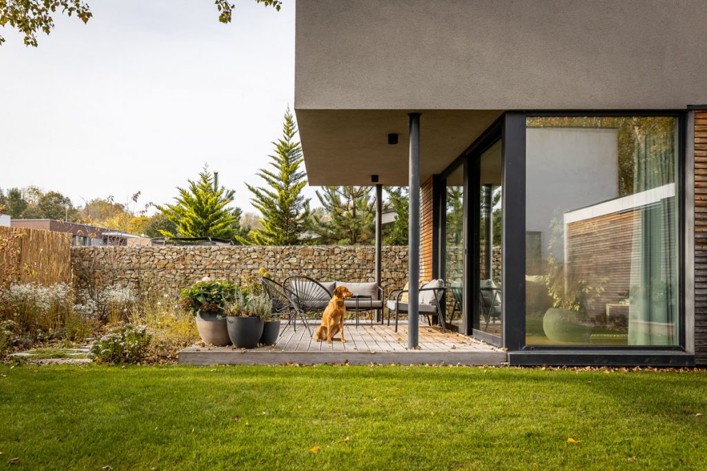 Moderný rodinný dom s terasou so sedením a kvetinami, gabionovým plotom a nenáročnou prírodnou záhradou.
