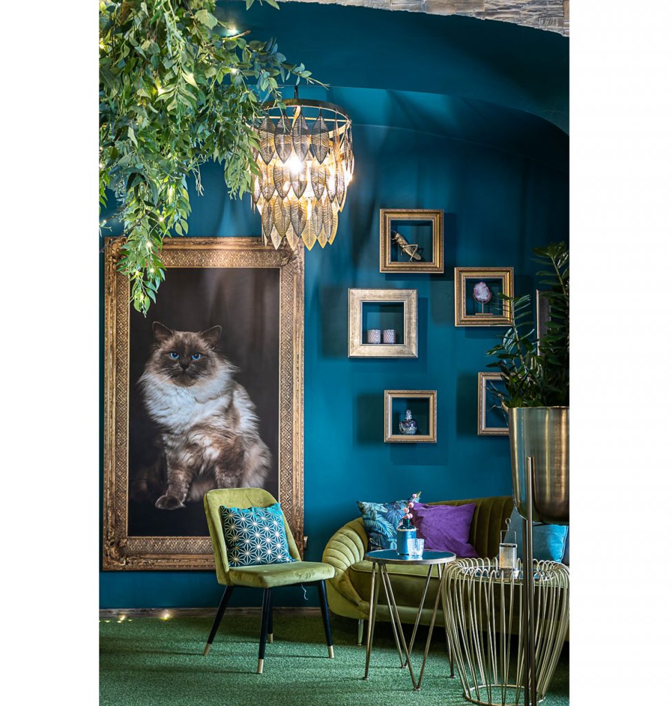 Kaviareň Ragdoll café: Detail interiéru s veľkým obrazom ragdoll mačky v mohutnom ráme, s glamour sedením v zelenom odtieni, kovovým dekoratívnym stolíkom a obrazovými rámami, ktoré slúžia ako police na dekorácie