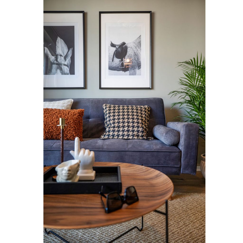 detail obývačky v pánskom štýle s farebnou čalúnenou sedačkou, s vankúšmi v škoricovej farbe a so vzorom kohútej stopy, s okrúhlym stolíkom a obrazmi s motívom koní
