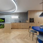 minimalistický industriálny interiér kuchyne spojenej s obývačkou s dizajnovým osvetlením v tvare kruhu a exteriérovým svietidlom nad kuchynskou linkou, dreveným kuchynským nábytkom, modrým dizajnovým stolom s kovovými stoličkami a betónovou stenou