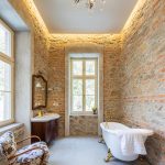 kúpeľňa s priznanými kamennými a tehlovými stenami, podsvieteným stropom, bielou vaňou na zlatých nožičkách, kvetinovým kreslom a starožitnou skrinkou s umývadlom