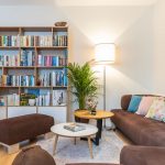 obývačka s hnedou čalúnenou sedačkou, okrúhlymi stolíkmi, závesnou skrinkou v lesklej bielej farbe a knižnicou