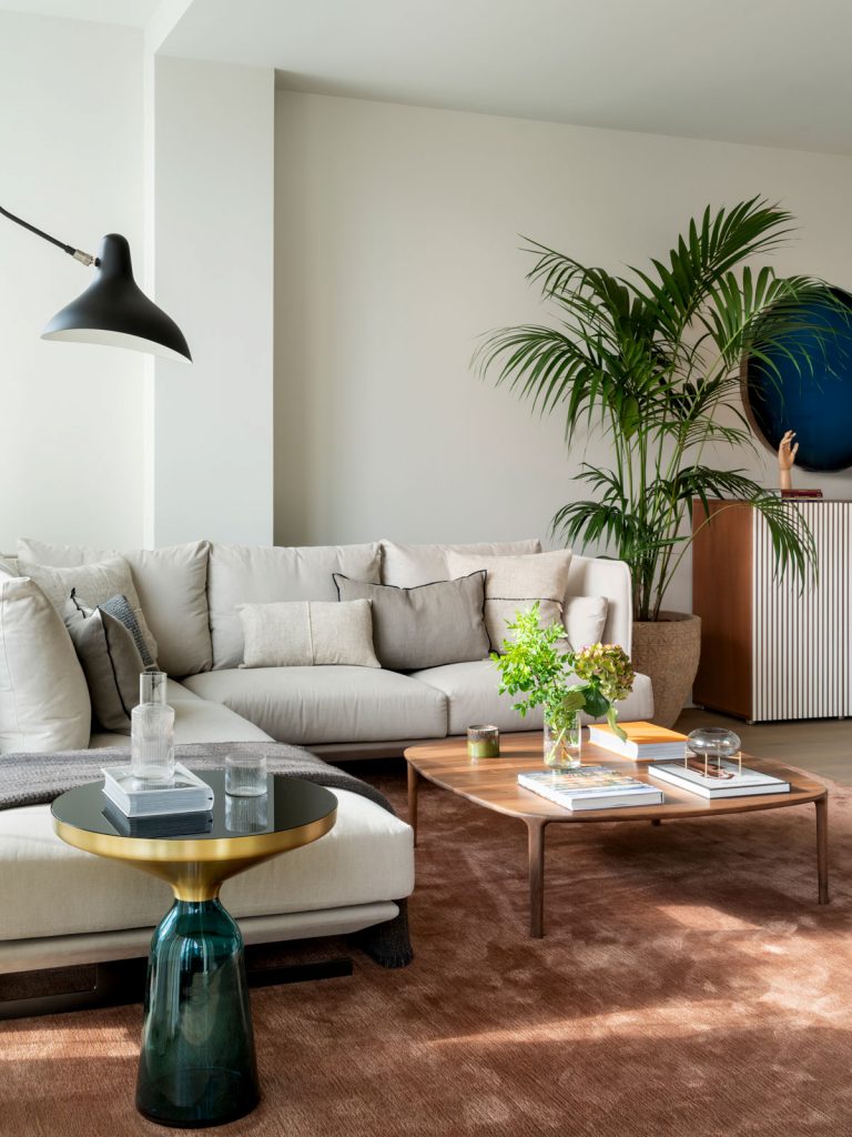 obývačka v zemitých tónoch, s krémovou sedačkou, hnedým kobercom a preskleným modrým okrúhlym stolíkom v tvare presýpacích hodín