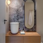 kúpeľňa so svetlou drevenou skrinkou, na ktorej je vysoké biele umývadlo a so stenou s abstraktnou maľovkou a oválnym zrkadlom