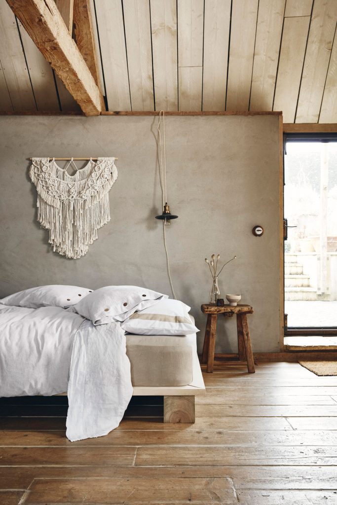 spálňa v neutrálnych odtieňoch, s betónovou stenou, závesnou makramé dekoráciou, drevenou posteľou a bielymi obliečkami