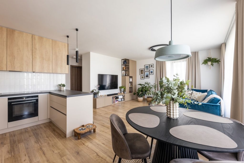 otvorený priestor kuchyne a obývačky s francúzskymi oknami, zariadený v štýle cozy living, s jednoduchou kuchyňou v drevodekore a bielej farbe, s jedálenský okrúhlym čiernym stolom, modrou sedačkou a uzatvoreným a otvoreným odkladacím priestorom v okolí televízie