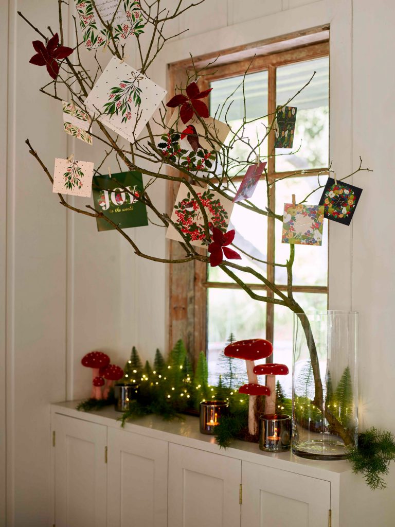 Sviatočné zátišie na parapete so sklenenou vázou s konárom, na ktorom sú povešané vianočné pohľadnice, vedľa vázy je dekorácia z muchotrávok