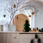 sviatočná sklenená váza s vetvičkou, na ktorej visia papierové ozdoby a papierové dekorácie vianočných stromčekov