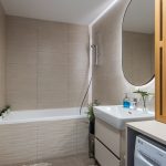 Kúpeľňa s béžovým obkladom, rastlinami, oválnym podsvieteným zrkadlom a lamelovou skrinkou