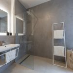 moderná kúpeľňa v apartmáne, so svetlou podlahou, betónovou stierkou, sprchovým kútom, podsvieteným zrkadlom a umývadlom s úložným priestorom