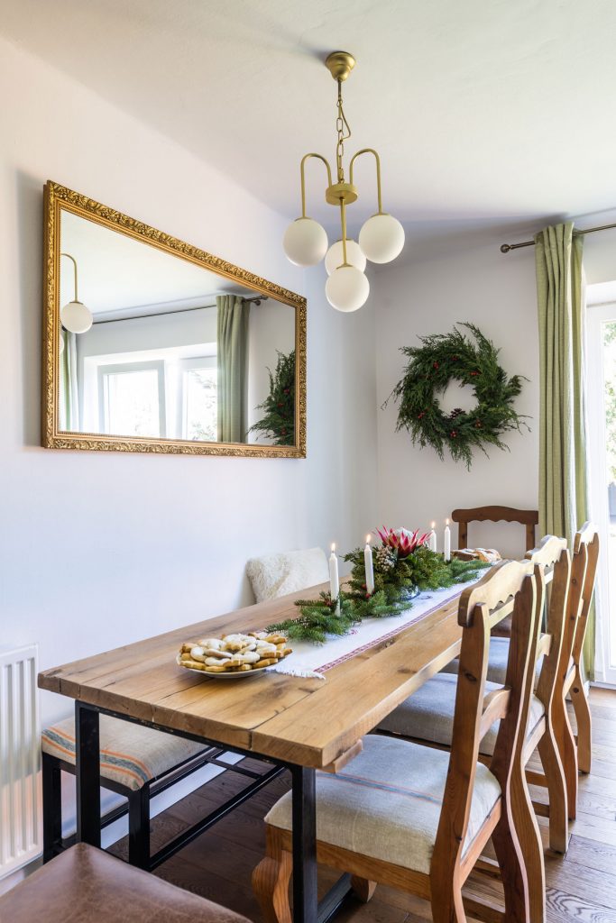 vianočne prestretý stôl vyrobený zo starých trámov, s behúňom, ihličnatými vetvičkami, drevenými stoličkami, zlatým retro lustrom, nad stolom visí zrkadlo v starožitnom ráme a veniec z čečiny