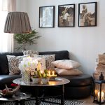 škandinávska vidiecka obývačka s čiernou koženou sedačkou a vankúšmi z prírodných materiálov, s konferenčným stolíkom s vianočným aranžmánom, geometrickým kobercom, textilným svietidlom a lampášom na podlahe