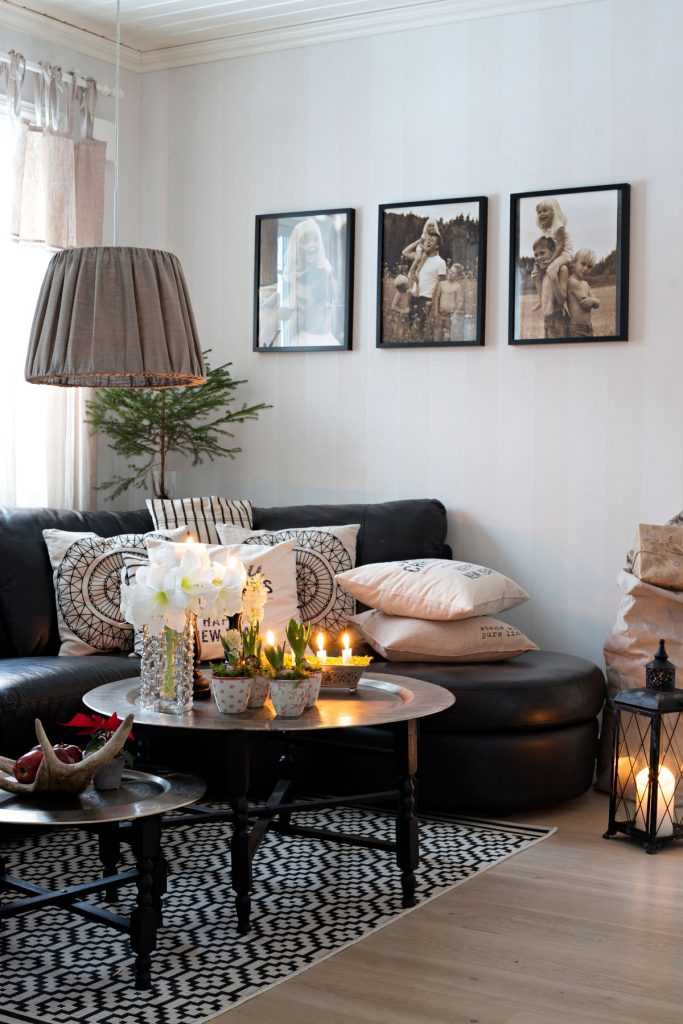 škandinávska vidiecka obývačka s čiernou koženou sedačkou a vankúšmi z prírodných materiálov, s konferenčným stolíkom s vianočným aranžmánom, geometrickým kobercom, textilným svietidlom a lampášom na podlahe
