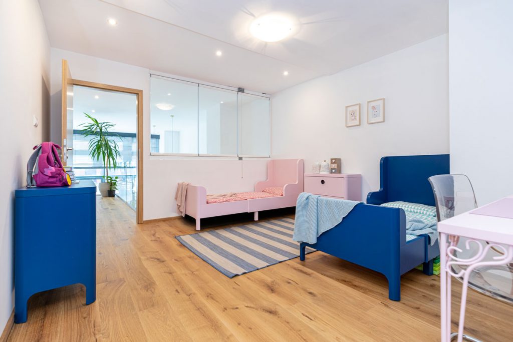 detská izba pre chlapca a dievča, s presklenou časťou steny a s jednoduchým zariadením v podobe modrej a ružovej postele, ktoré rastú spolu s deťmi, modrej komody a ružového pracovného stolíka