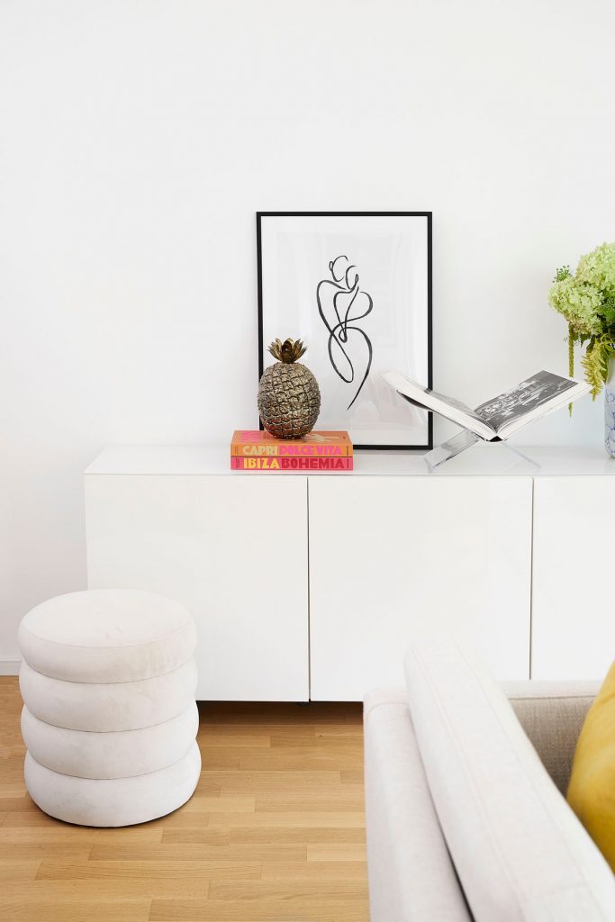 biela skrinka, dizajnový biely taburet, na skrinke stojí dekorácia zlatého ananásu a abstraktný obrázok