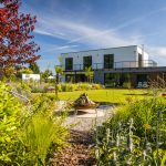 Pohľad na rodinný dom a okrasnú modernú vidiecku záhradu s trávnikom, záhonmi a posedením s prenosným ohniskom