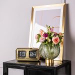 Čierna presklená skrinka so zlatým zrkadlom, retro digitálnymi hodinami a zelenou vintage vázou s kvetmi