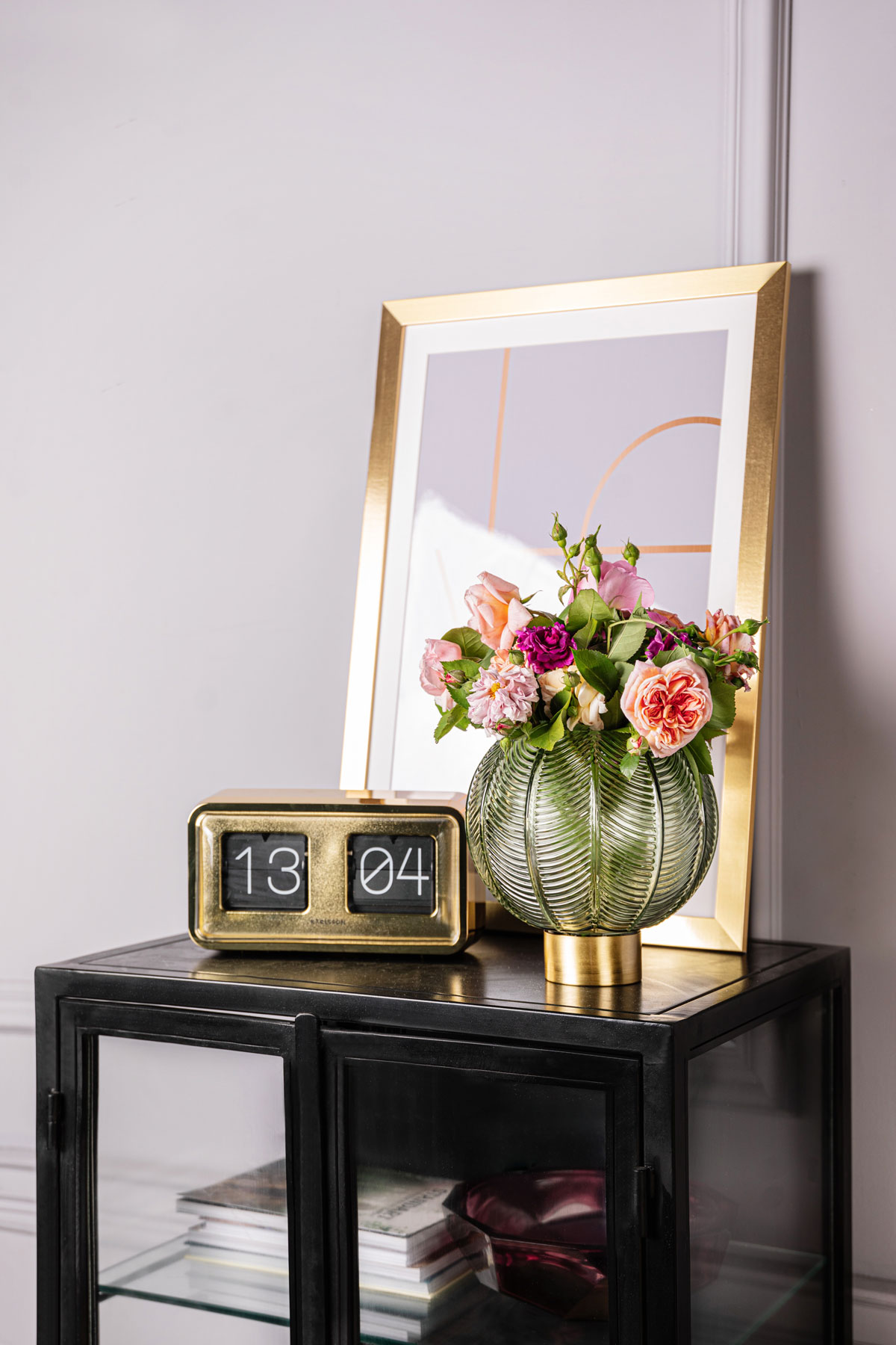 Čierna presklená skrinka so zlatým zrkadlom, retro digitálnymi hodinami a zelenou vintage vázou s kvetmi