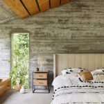 Spálňa s betónovou stenou, posteľou s výrazným koženým čelom a etno posteľným prádlom a drevenou lavicou s odkladacím priestorom pozdĺž okna