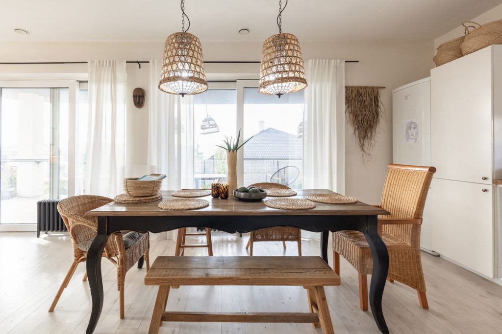 jedáleň v rodinnom dome s dreveným jedálenským stolom s ozdobnými nožičkami, drevenou lavicou na sedenie a rozdielnymi prútenými stoličkami a dvojicou prútených lustrov nad stolom