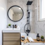 moderná kúpeľňa s vaňou, drevenou skrinkou s umývadlom, čiernym zrkadlom, čiernou sprchovacou hlavicou a batériami a obkladom s imitáciou mramoru na stene
