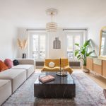 svetlý interiér modernej obývačky so smotanovou minimalistickou sedačkou s farebnými vankúšmi, mramorovým čiernym stolíkom, žltým kreslom a svetlou skrinkou na nožičkách, na ktorej stojí zrkadlo v zlatom ráme