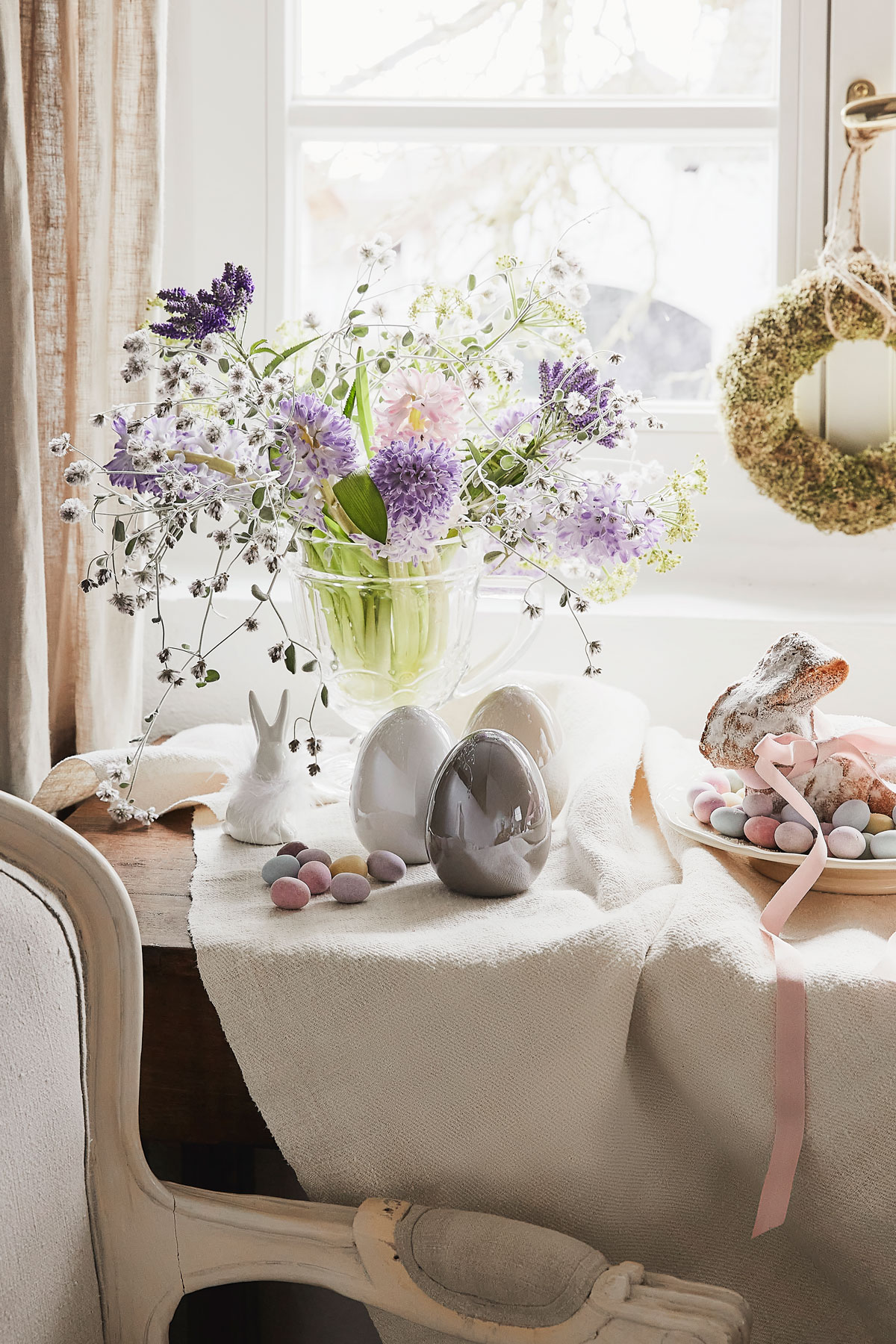 Aranžmán na Veľkú noc s prírodným vencom na okne, vázou s kvetmi, barančekom a keramickými vajíčkami a zajacom