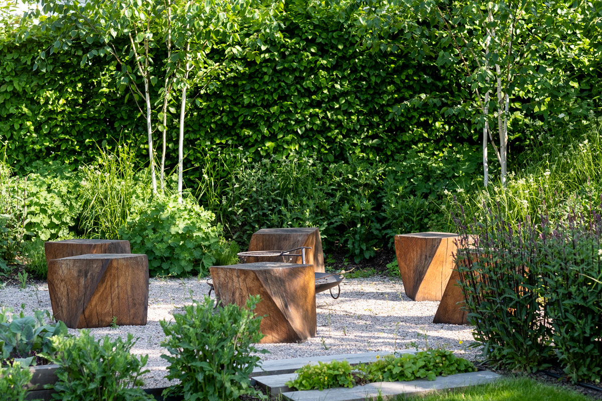 zóna s ohniskom v záhrade, vysypaná štrkom, obklopená výsadbami trvaliek, briez a krovín, ako sedenie sú použité drevené kocky od sochára Michala Sýkoru