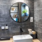 moderná kúpeľňa s obkladom vzhľadu kameňa, dubovou skrinkou s bielym umývadlom a čiernou batériou a okrúhlym zrkadlom v čiernom ráme