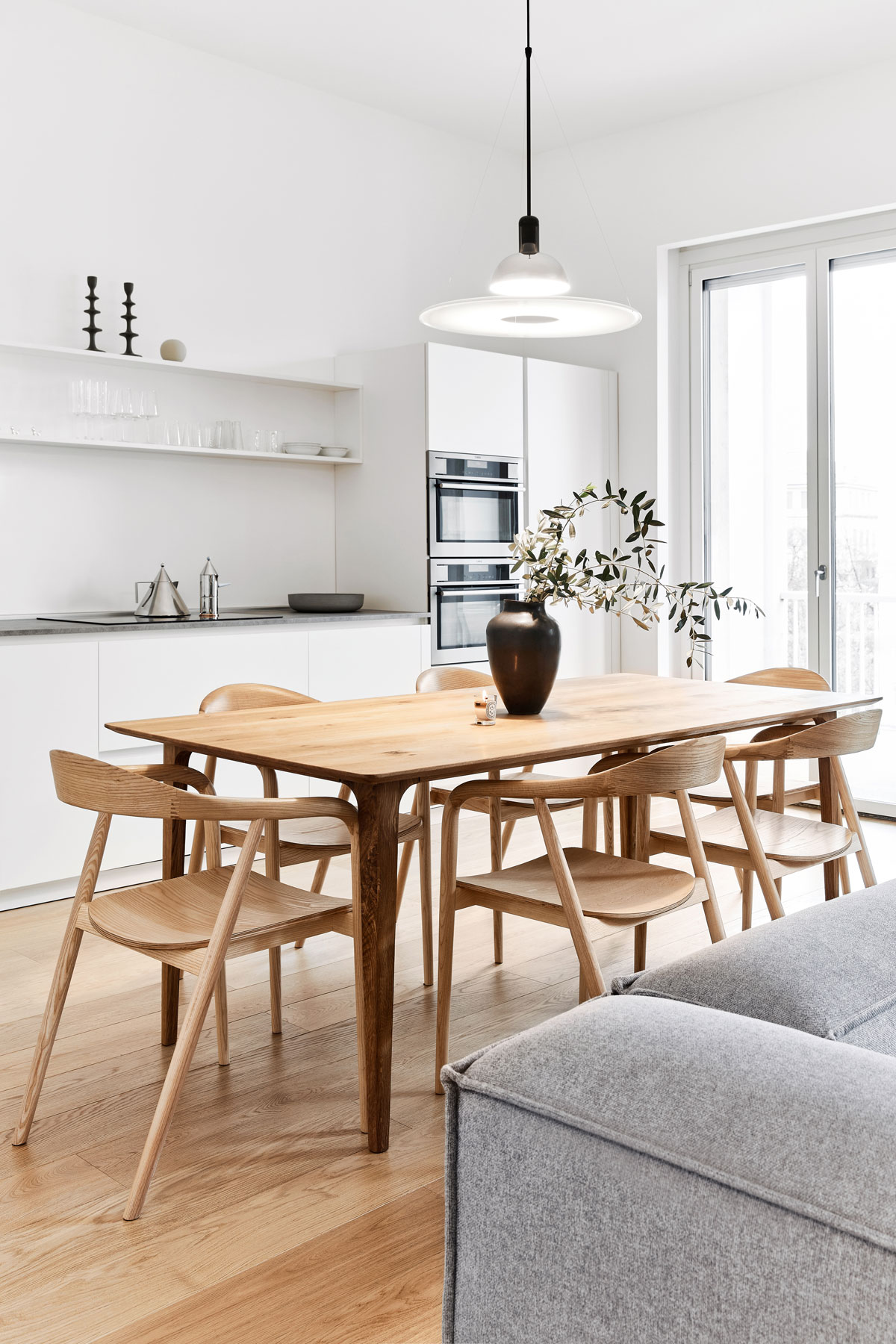 Biela kuchynská linka s uzavretým spodkom a otvorenými vrchnými policami, drevený jedálenský stôl v otvorenom koncepte s obývačkou