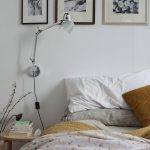 pohľad na posteľ v spálni s prírodnými ľanovými a bavlnenými obliečkami v tlmenej palete farieb, vedľa postele je jednoduchý drevený stolík, nad posteľou je umiestnená sivá kovová lampa a čiernobiele obrazy