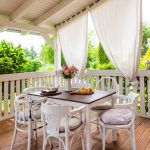 drevená terasa so zábradlím a závesmi a bielym vidieckym stolom so stoličkami
