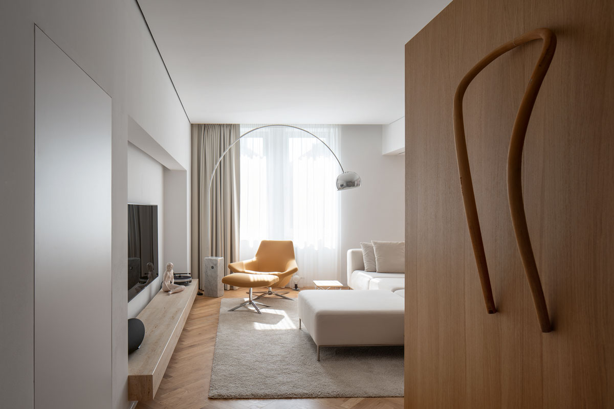 Obývačka so smotanovou sedačkou, žltým dizajnovým kreslom, svetlým kobercom, televízia je vsadená do výklenku v stene spolu s drevenou masívnou policou