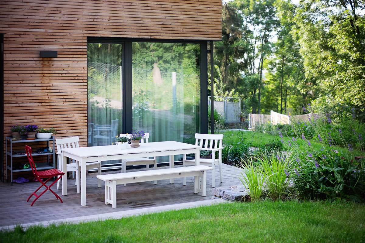 Moderná novostavba rodinného domu s dreveným obkladom, pred domov je terasa s bielym záhradným nábytkom, okolo domu je okrasná záhrada susediaca s lesom