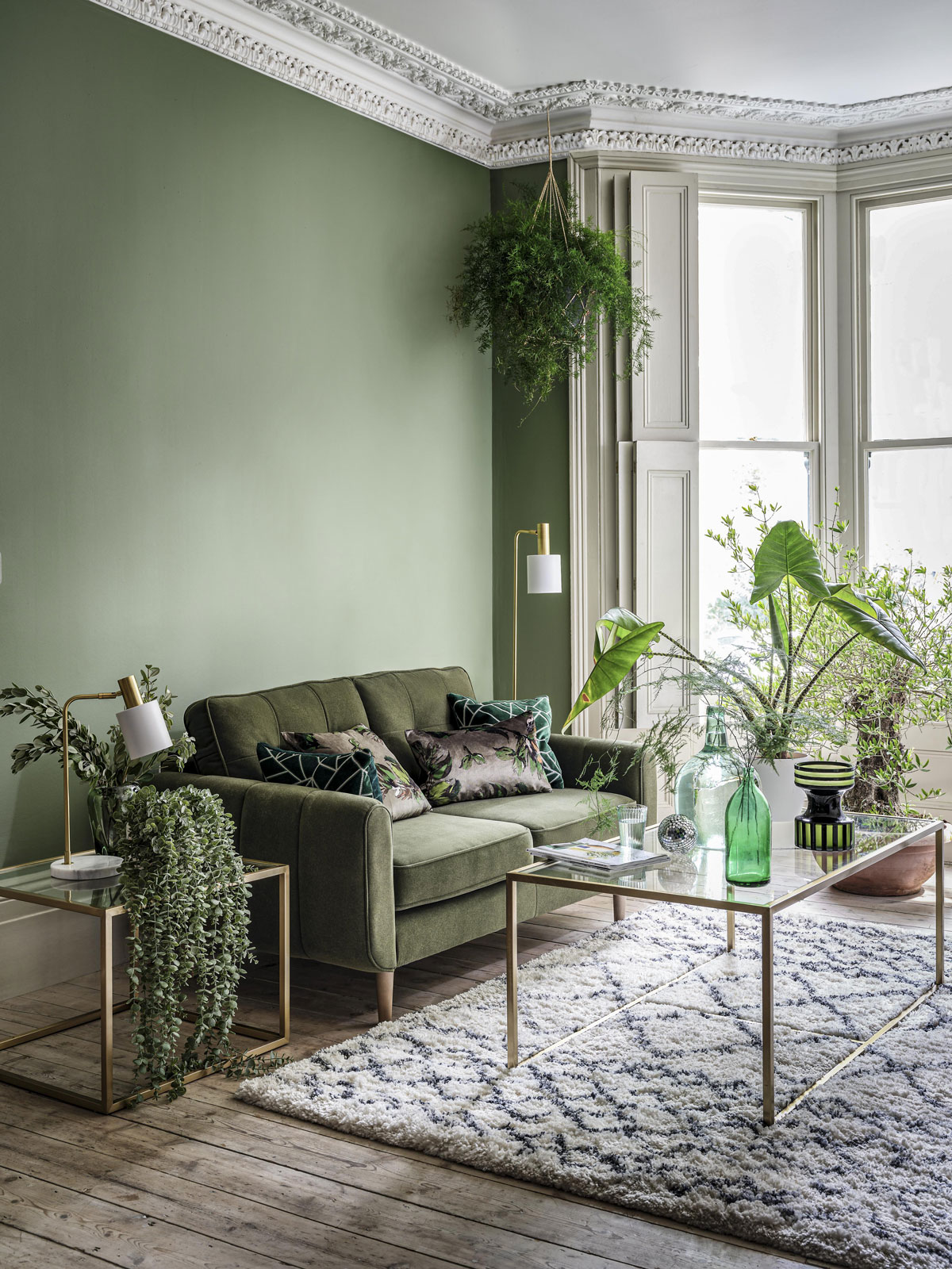Interiér obývačky ladenej do zelena. Obývačka má vysoké zdobené stropy, zelenú stenu, pri ktorej stojí zamatová olivová sedačka, pri sedačke sú kovové stolíky so sklom, v miestnosti sú zelené izbové rastliny