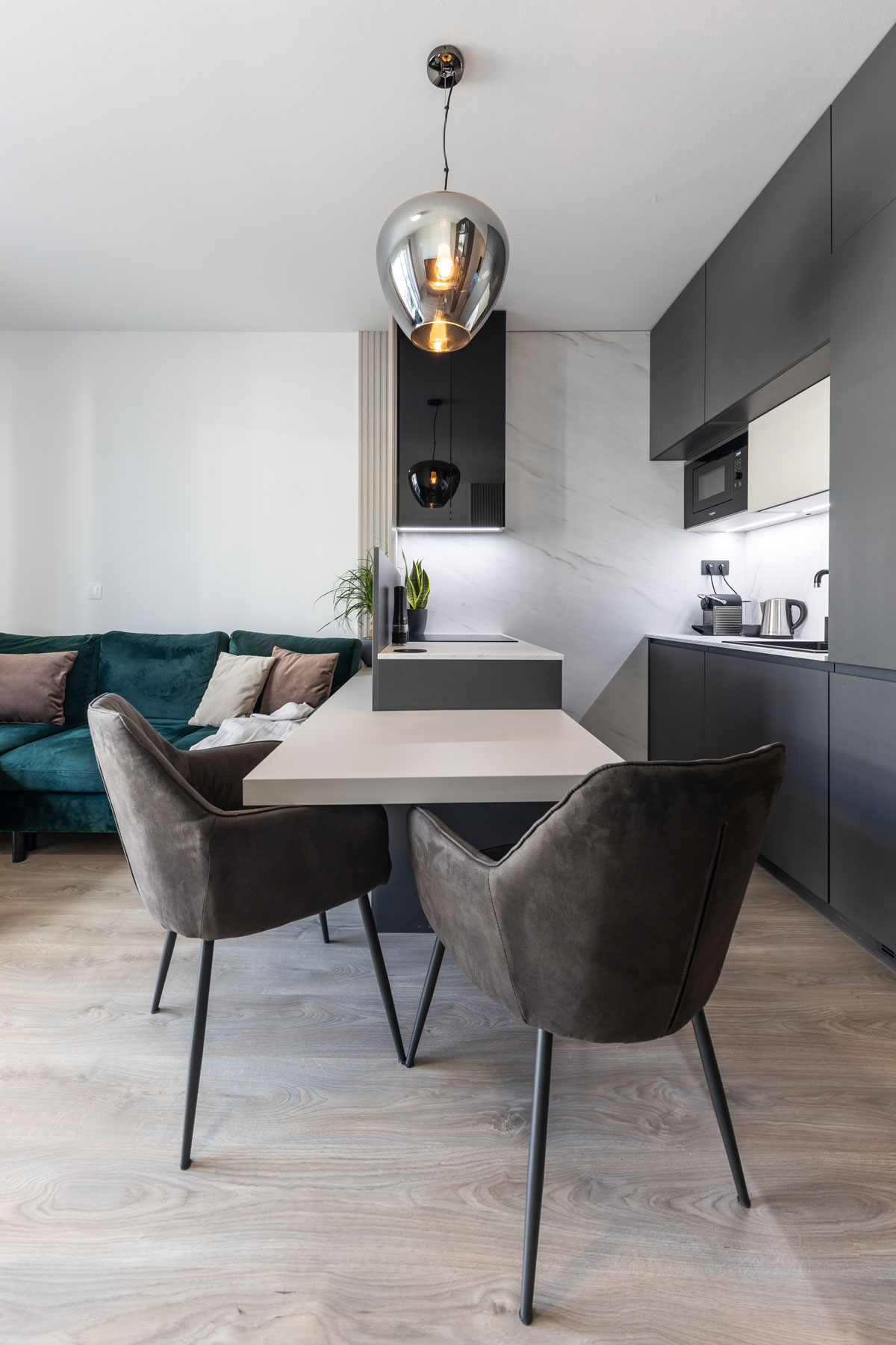 moderný malý byt s minimalistickou kuchynskou linkou v tmavej farbe, oproti je polostrov s čalúnenými stoličkami, ten oddeľuje kuchyňu od obývačky s tmavozelenou sedačkou