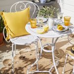 Biely kovový stolík s bielou kovovou stoličkou, dopĺňa ich žltý vankúš a žlté riady na stolíku