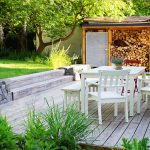 drevená terasa s bielym záhradným nábytkom, za terasou je drevená stavba na uskladnenie dreva, od terasy vedú schodíky na o úroveň vyššiu trávnatú plochu