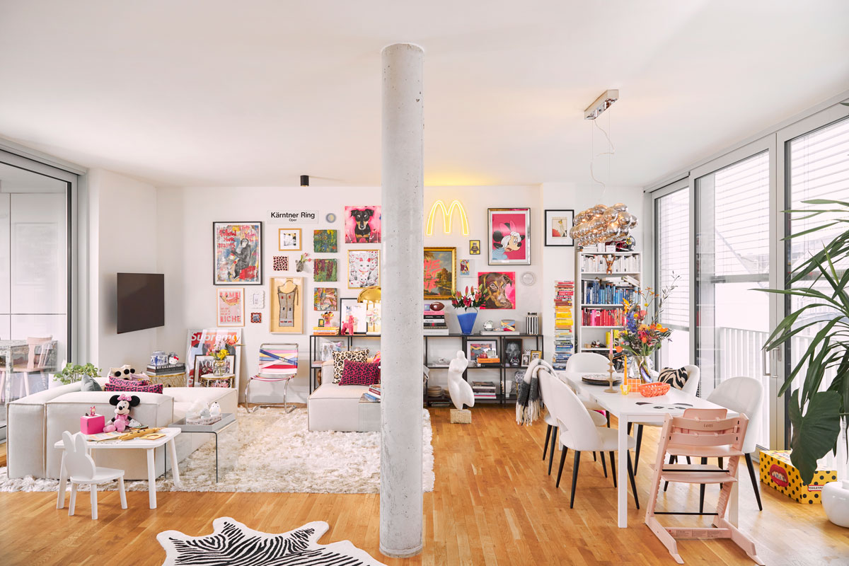 otvorený priestor jedálne a obývačky v maximalistickom apartmáne Mariny Hoermanseder, jeho základom je biela farba na jedálenskom stole, stoličkách a sedačke s kreslom, priestor oživujú rôzne extravagantné farebné dekorácie a doplnky, knihy a moderné obrazy tvoriace galériu